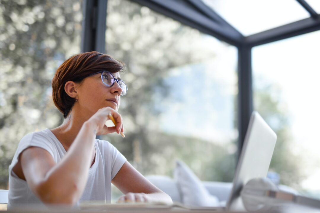 Woman working at laptop in front of large glass walls with foliage outside. Kvinna jobbar vid laptop framför stora glasväggar med lövverk utanför.
