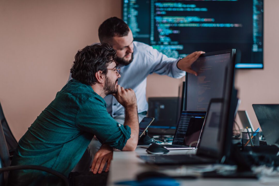 Two developers at a desk with screens showing the NOC Network Operations Centre and IT support. Två utvecklare vid ett skrivbord med skärmar som visar NOC Network Operations Center och IT-support.