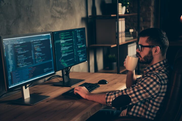A coder with a coffee cup at computer screens. En kodare med kaffekopp vid datorskärmar.